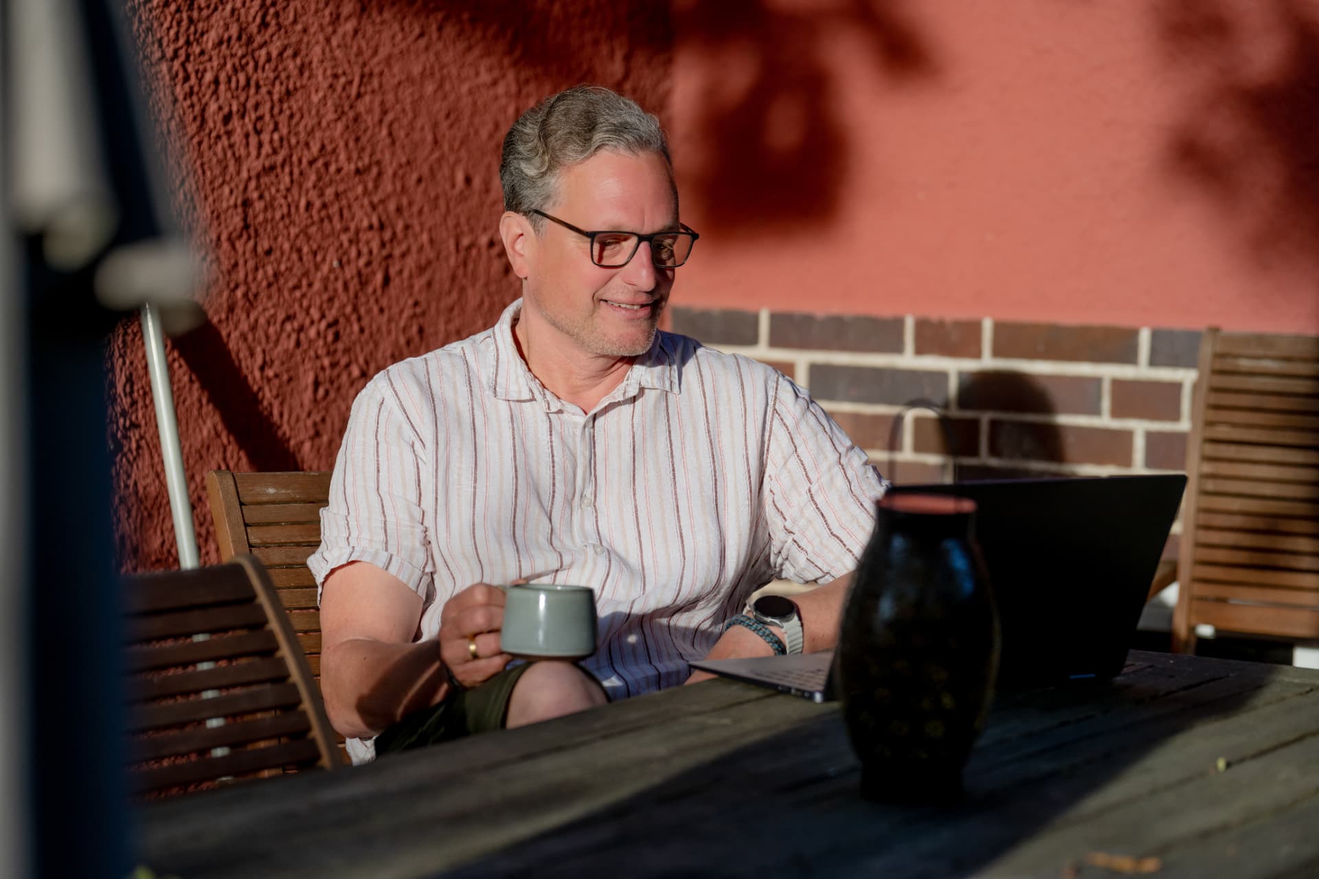 Lars Schwichtenberg at a wooden table outdoors, a mug in his hand, working on a laptop